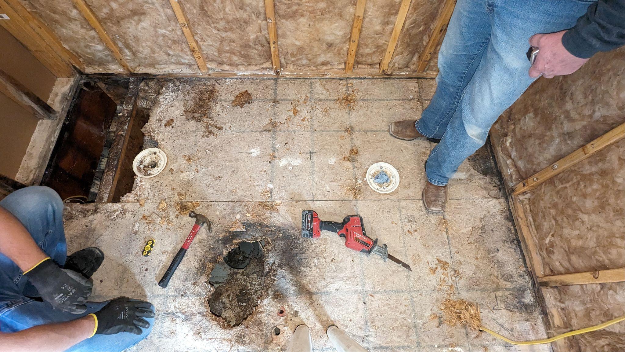 Flooring repair in progress, showing a damaged area with tools like a hammer and drill. Two individuals are working on the project.